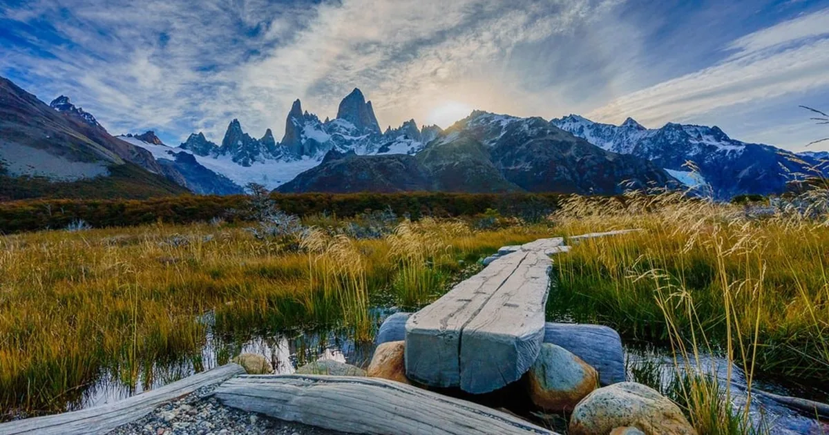 A wooden boardwalk across a golden meadow towards the snowy Fitz Roy massif