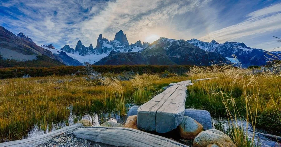 A wooden boardwalk across a golden meadow towards the snowy Fitz Roy massif on a clear day