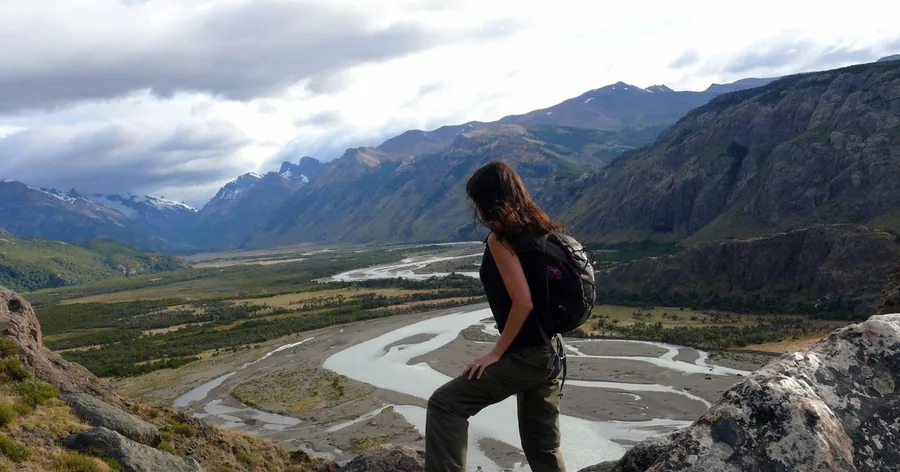 A hiker on a rocky ledge looking down the braided Río de las Vueltas valley towards distant mountains
