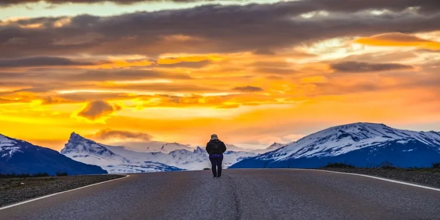 A lone walker on a straight road at sunset with snowy peaks on the horizon