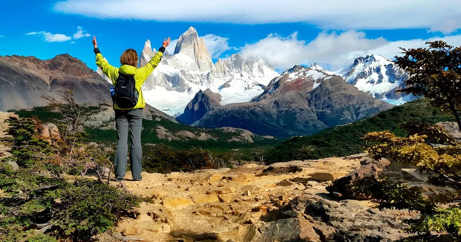 A hiker with arms raised on a rocky viewpoint facing the granite spires of Fitz Roy